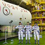 In the Integration Facility at the Baikonur Cosmodrome in Kazakhstan, Expedition 44 crew members Kjell Lindgren of NASA (left), Oleg Kononenko of the Russian Federal Space Agency (Roscosmos, center) and Kimiya Yui of the Japan Aerospace Exploration Agency (right) wave to reporters by the upper stage of their Soyuz booster rocket July 11 prior to a fit check dress rehearsal session. The trio will launch July 23, Kazakh time from Baikonur in their Soyuz TMA-17M spacecraft for a five-month mission on the International Space Station. Credit: Gagarin Cosmonaut Training Center